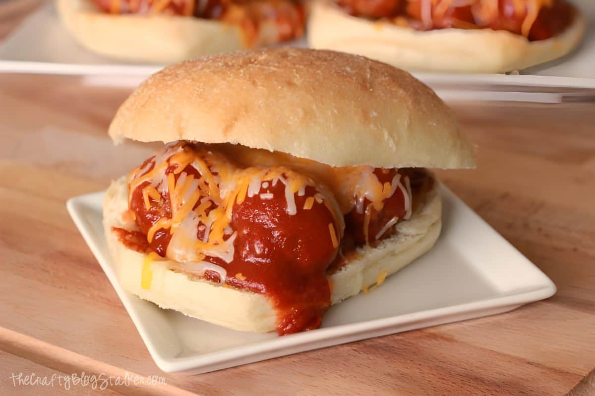 A meatball sandwich with tomato sauce and shredded cheese on a white square plate, with another sandwich in the background.