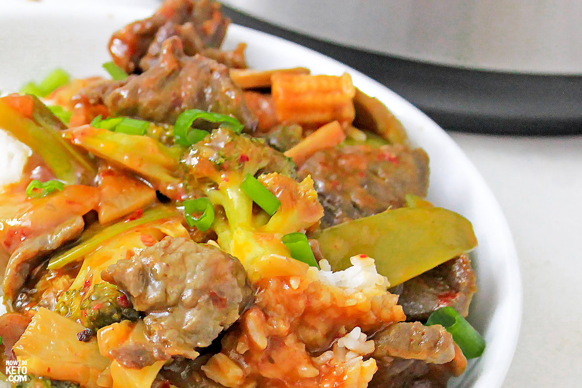 A close-up of a plate with beef stir-fry, mixed vegetables, and rice, garnished with chopped green onions.