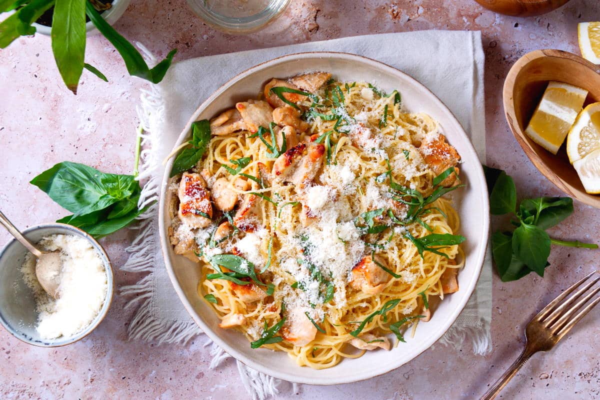 A bowl of creamy pasta with grilled chicken, garnished with grated cheese and fresh herbs, sits on a cloth napkin, surrounded by basil leaves and lemon wedges.