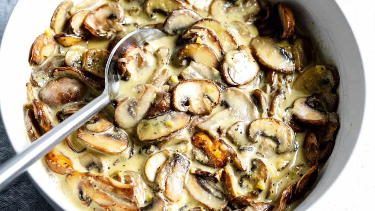 A bowl of creamy cooked mushrooms with a spoon, featuring sliced mushrooms in a thick, beige sauce.