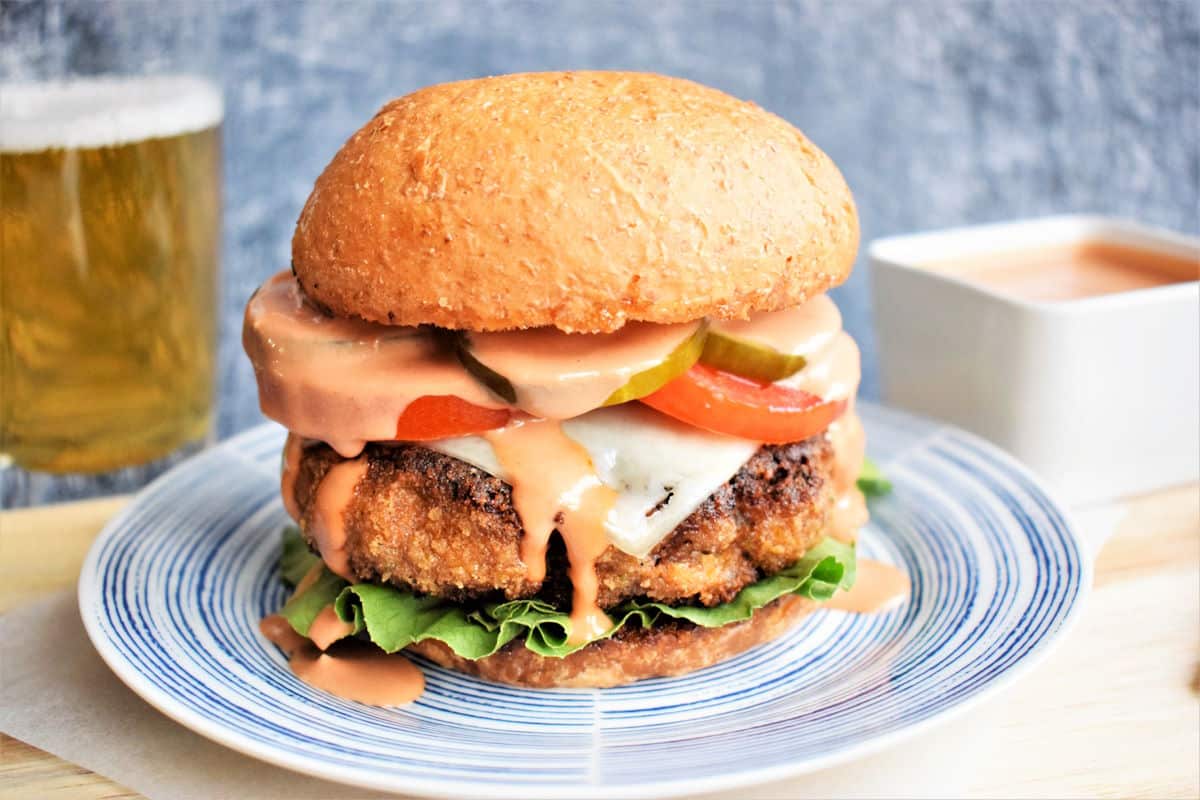 A breaded veggie burger with lettuce, tomato, pickles, cheese, and sauce on a wheat bun, served on a plate with a drink and extra sauce in the background.