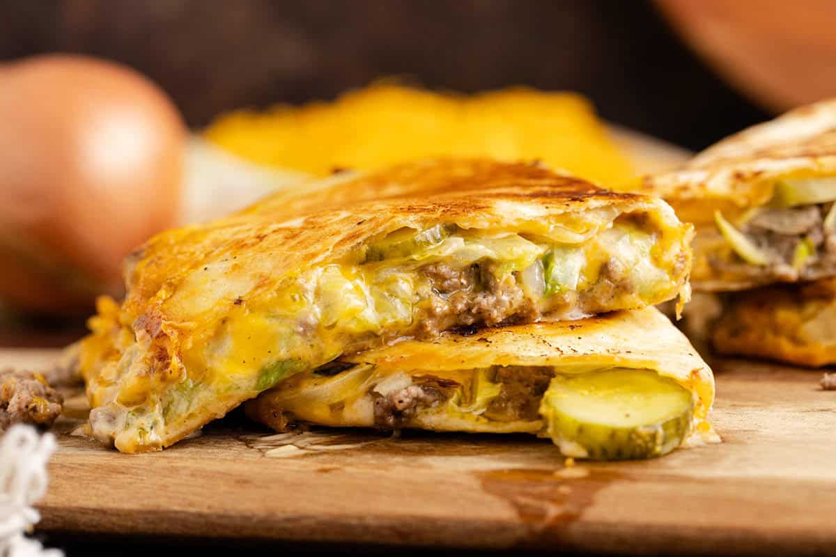 A close-up of a quesadilla cut in half, filled with ground beef, melted cheese, pickles, and onions, displayed on a wooden board.