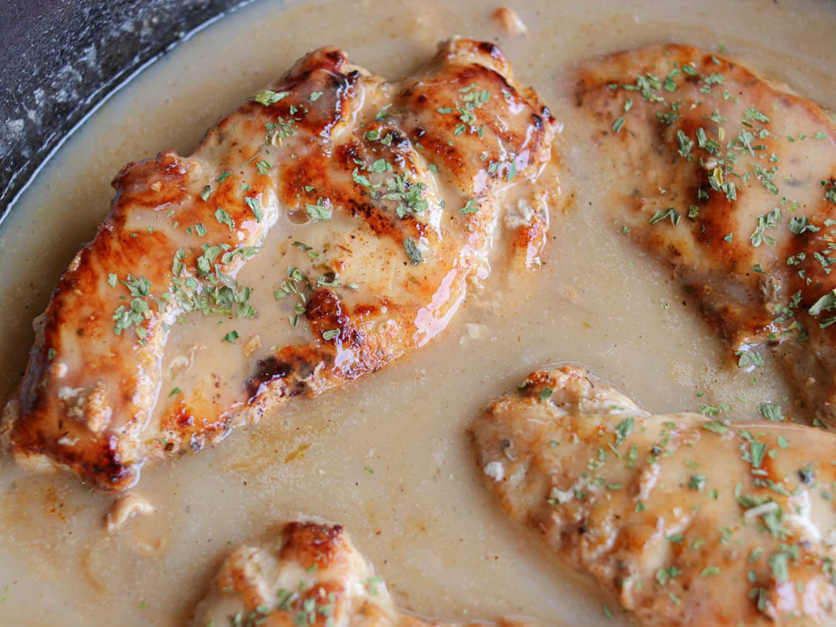 Three cooked chicken breasts covered in a creamy sauce and sprinkled with chopped herbs in a pan.