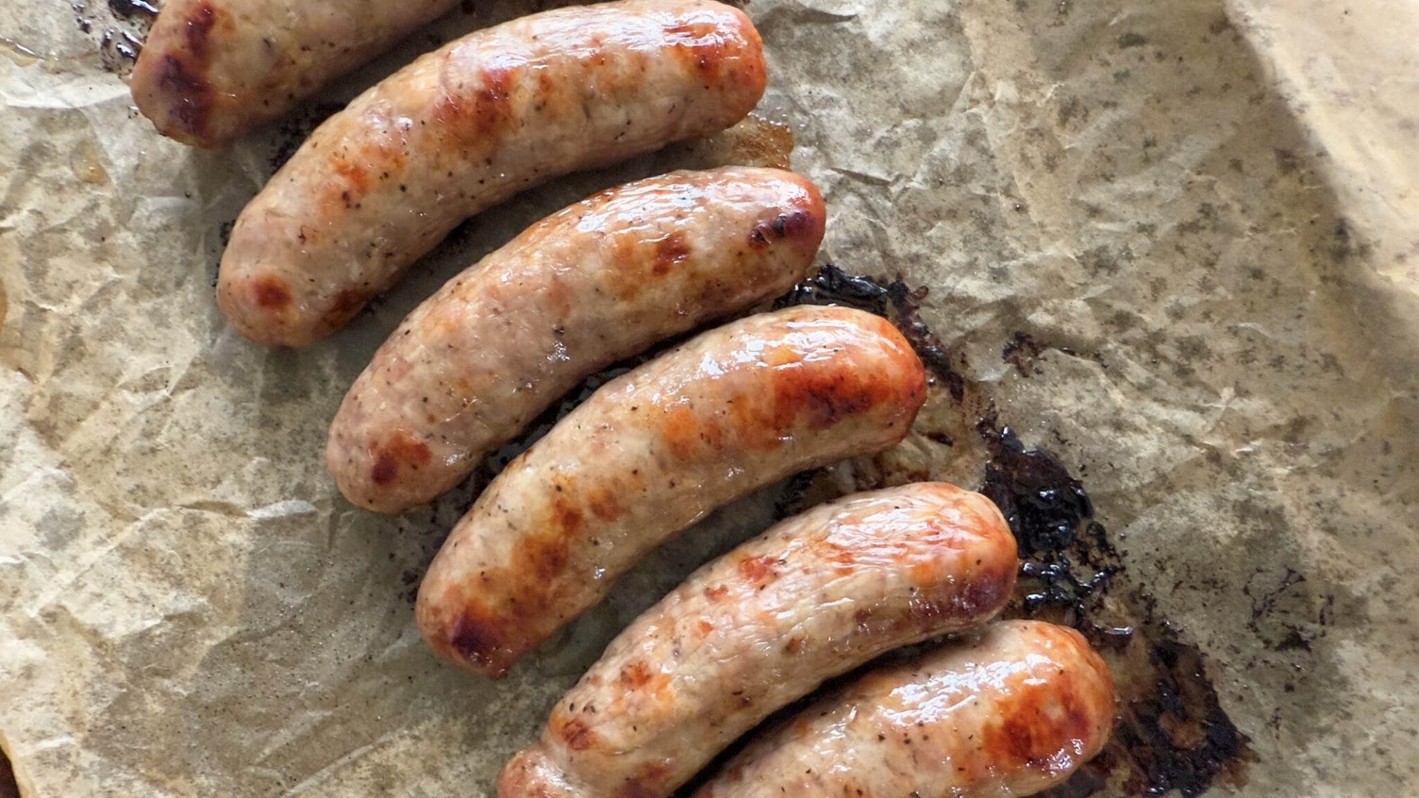 Six cooked sausages arranged in a row on a piece of brown parchment paper.