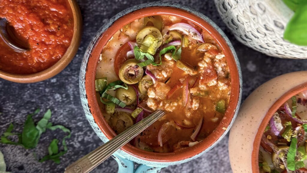 A bowl of tomato-based soup with sliced olives, red onions, herbs, and ground meat, with a spoon inside. A side dish of red sauce and fresh herbs appear nearby.