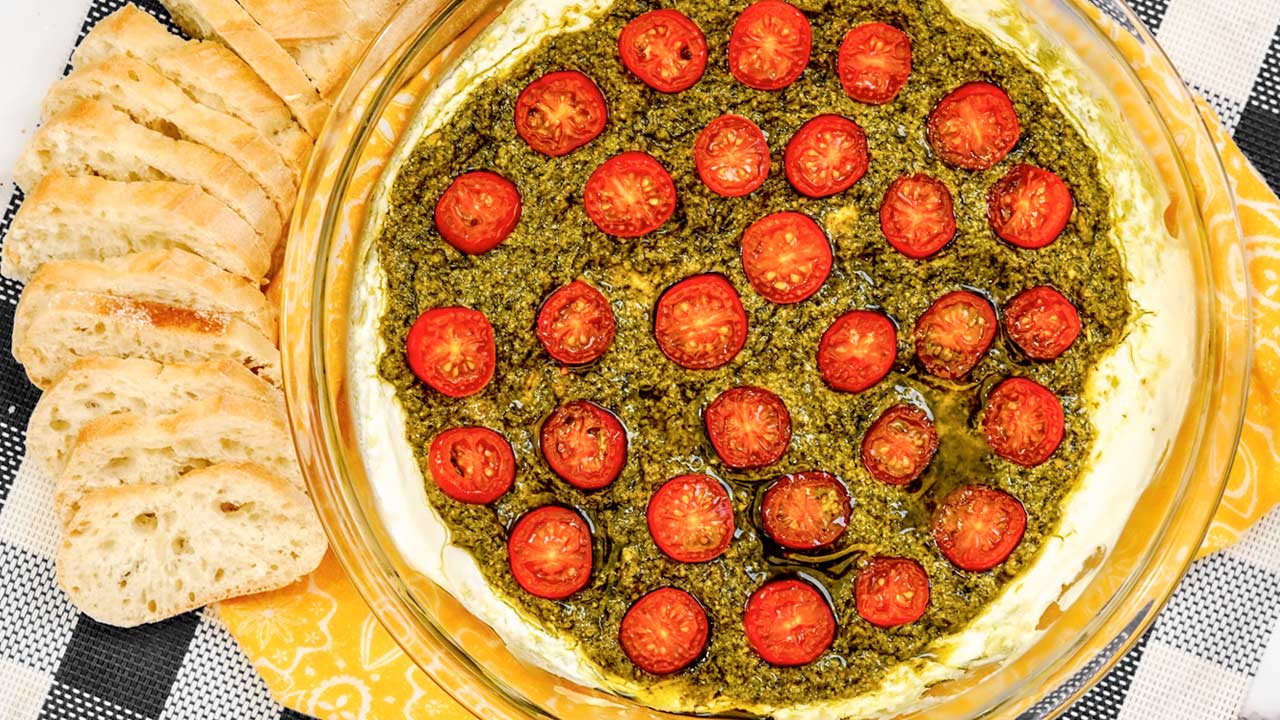 A round glass dish with a dip layered with green pesto, sliced cherry tomatoes, and surrounded by slices of bread on a checkered cloth.