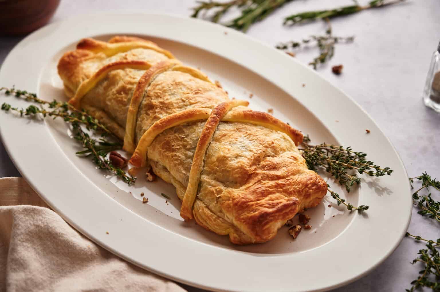 A golden-brown puff pastry roll sits on a white oval plate, garnished with sprigs of fresh thyme.