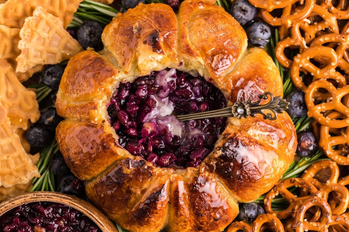 A baked pastry with a glossy, golden-brown crust is filled with dark berry compote. Surrounded by blueberries, pretzels, and waffle cookies, a decorative spoon rests in the filling.