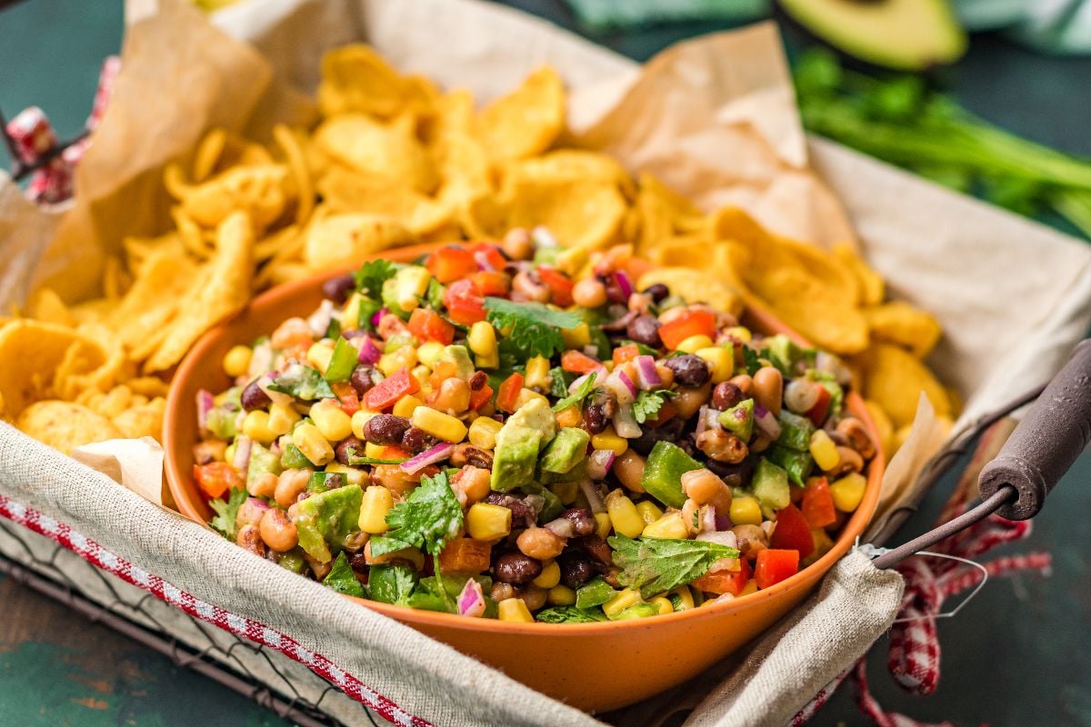 A bowl of colorful bean and corn salsa with diced vegetables, surrounded by corn chips in a tray lined with parchment paper.