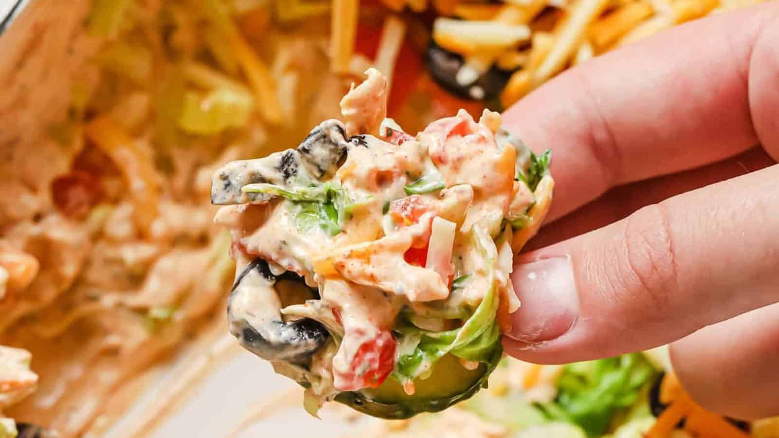 A hand holds a chip topped with a layered dip containing lettuce, olives, tomatoes, cheese, and a creamy sauce.