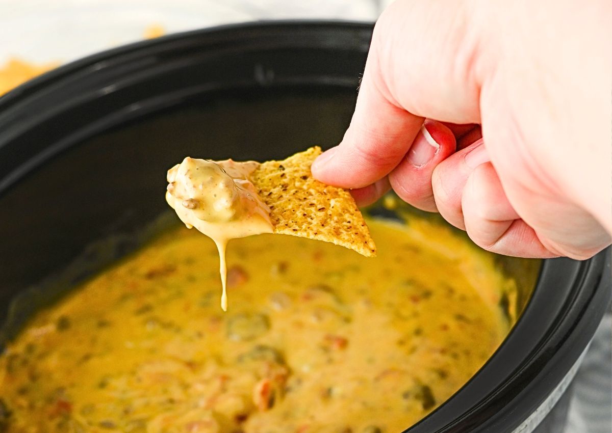 A hand dips a tortilla chip into a slow cooker filled with melted cheese dip containing visible bits of vegetables.