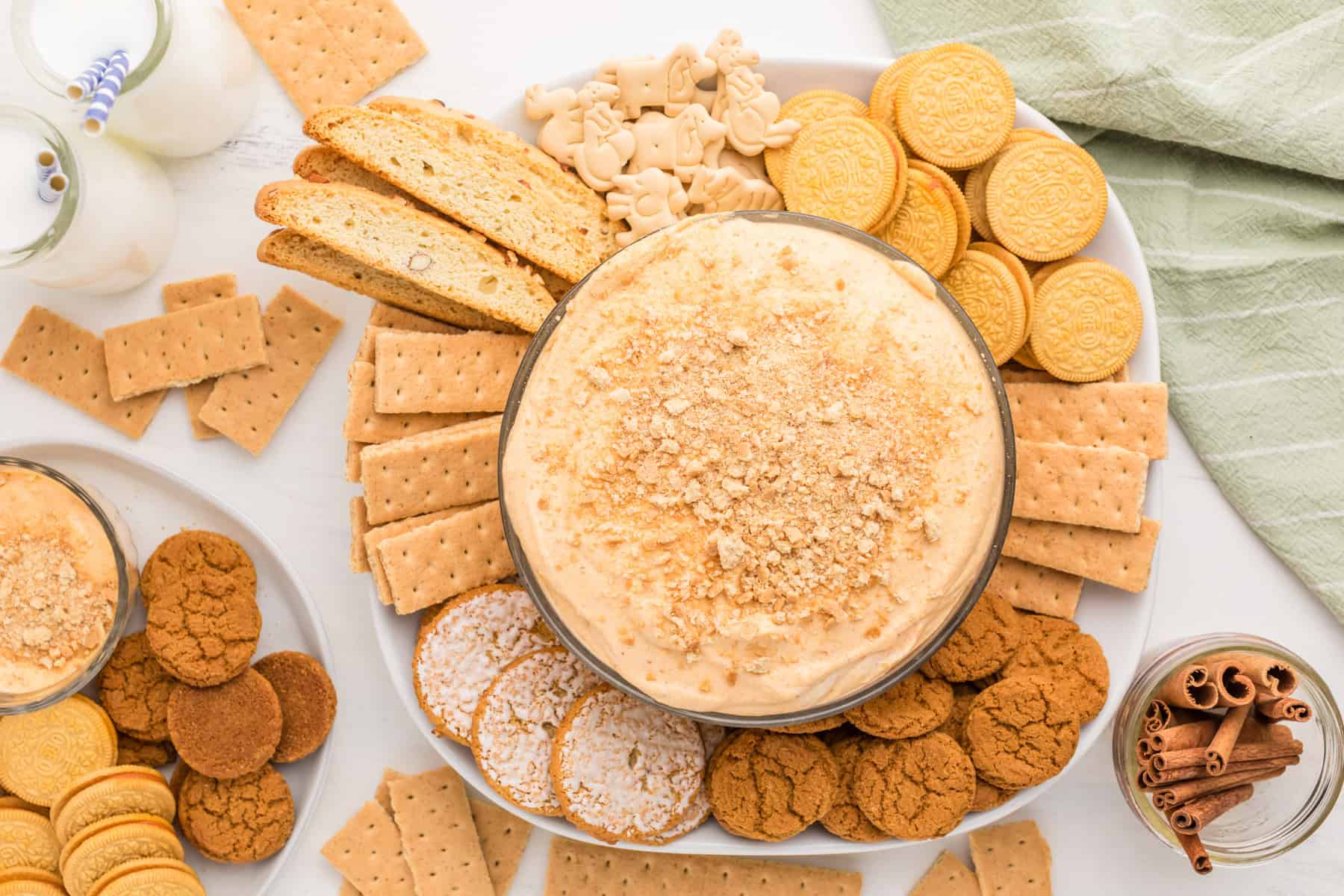 A platter with a bowl of creamy dip topped with crumbs, surrounded by various cookies, graham crackers, biscotti, and animal crackers, with glasses of milk nearby.