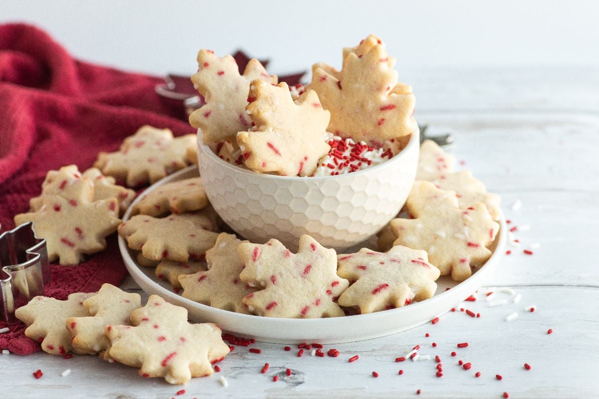 Plate and bowl filled with maple-leaf-shaped cookies topped with red sprinkles, set on a white wooden surface with a red cloth in the background.