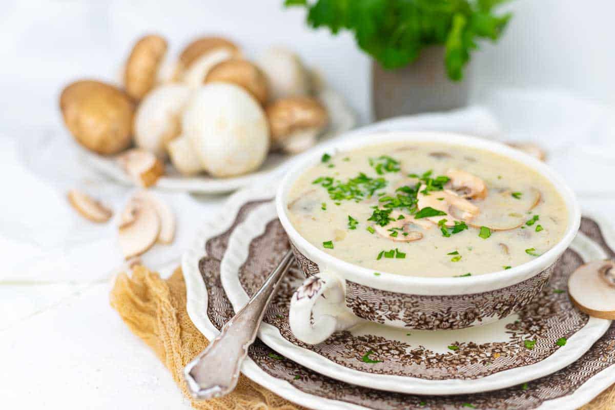 A bowl of creamy mushroom soup garnished with chopped herbs, placed on patterned dishes with a spoon, and fresh mushrooms and parsley in the background.