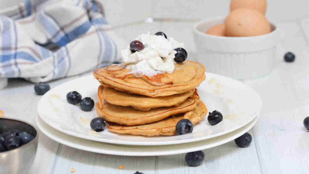 A stack of pancakes topped with whipped cream and blueberries sits on a white plate, with extra blueberries scattered around and eggs in a bowl in the background.