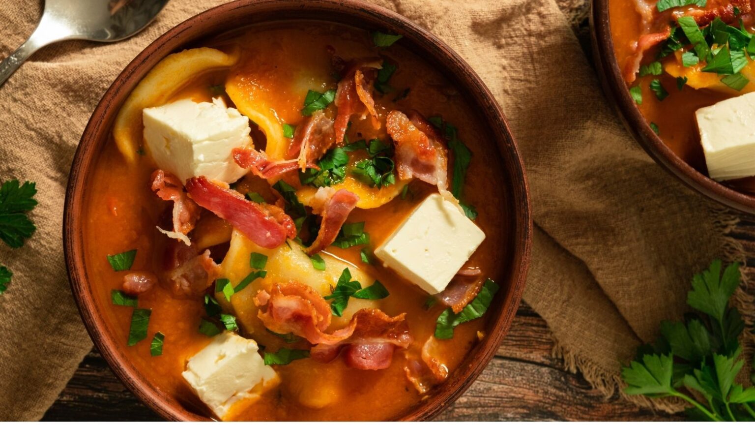 A bowl of soup with pasta, crispy bacon, cubes of white cheese, and chopped parsley on top, set on a brown cloth napkin.