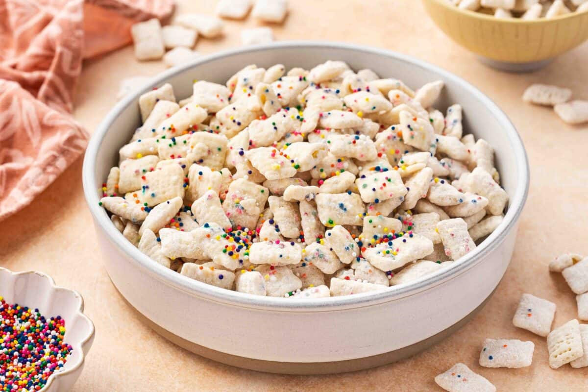 A bowl filled with powdered sugar-coated cereal pieces topped with colorful sprinkles, placed on a light surface with additional cereal and sprinkles nearby.