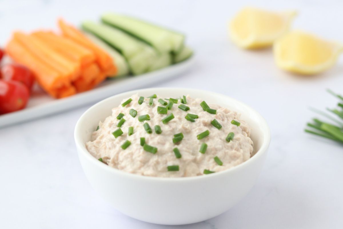 A white bowl of creamy dip topped with chopped chives sits in the foreground; sliced carrots, cucumbers, tomatoes, and lemon wedges are in the background.