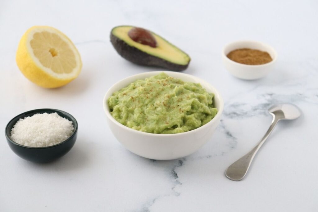 A bowl of guacamole is surrounded by half a lemon, half an avocado, a small bowl of salt, a small bowl of spices, and a spoon on a white surface.