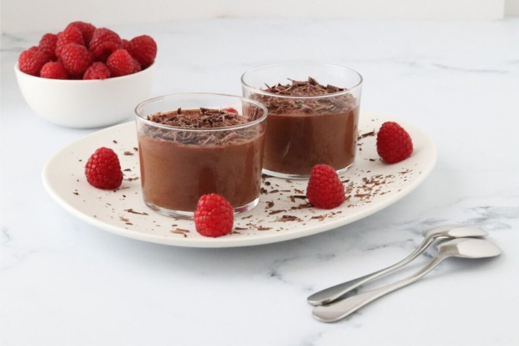 Two glasses of chocolate mousse topped with chocolate shavings, served on a white plate with fresh raspberries, with a bowl of raspberries and two spoons beside them.
