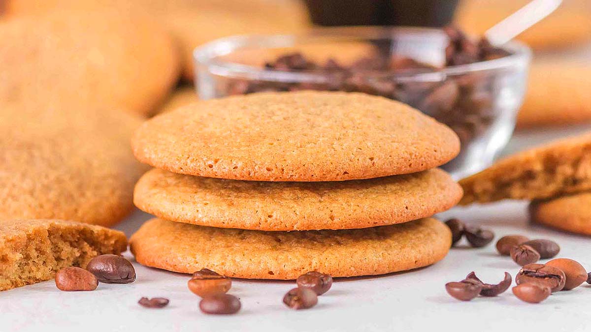 Three golden brown cookies are stacked on a surface with coffee beans scattered around and a glass bowl of coffee beans in the background.