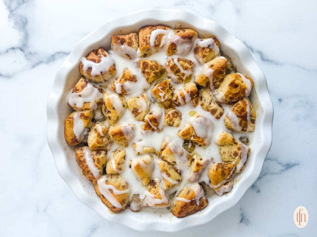 A white baking dish filled with baked cinnamon roll pieces topped with white icing, placed on a marble surface.