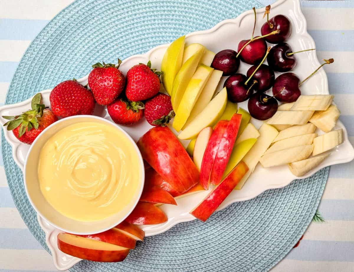 A platter with strawberries, apple slices, banana slices, cherries, and a bowl of creamy dip on a light blue placemat.