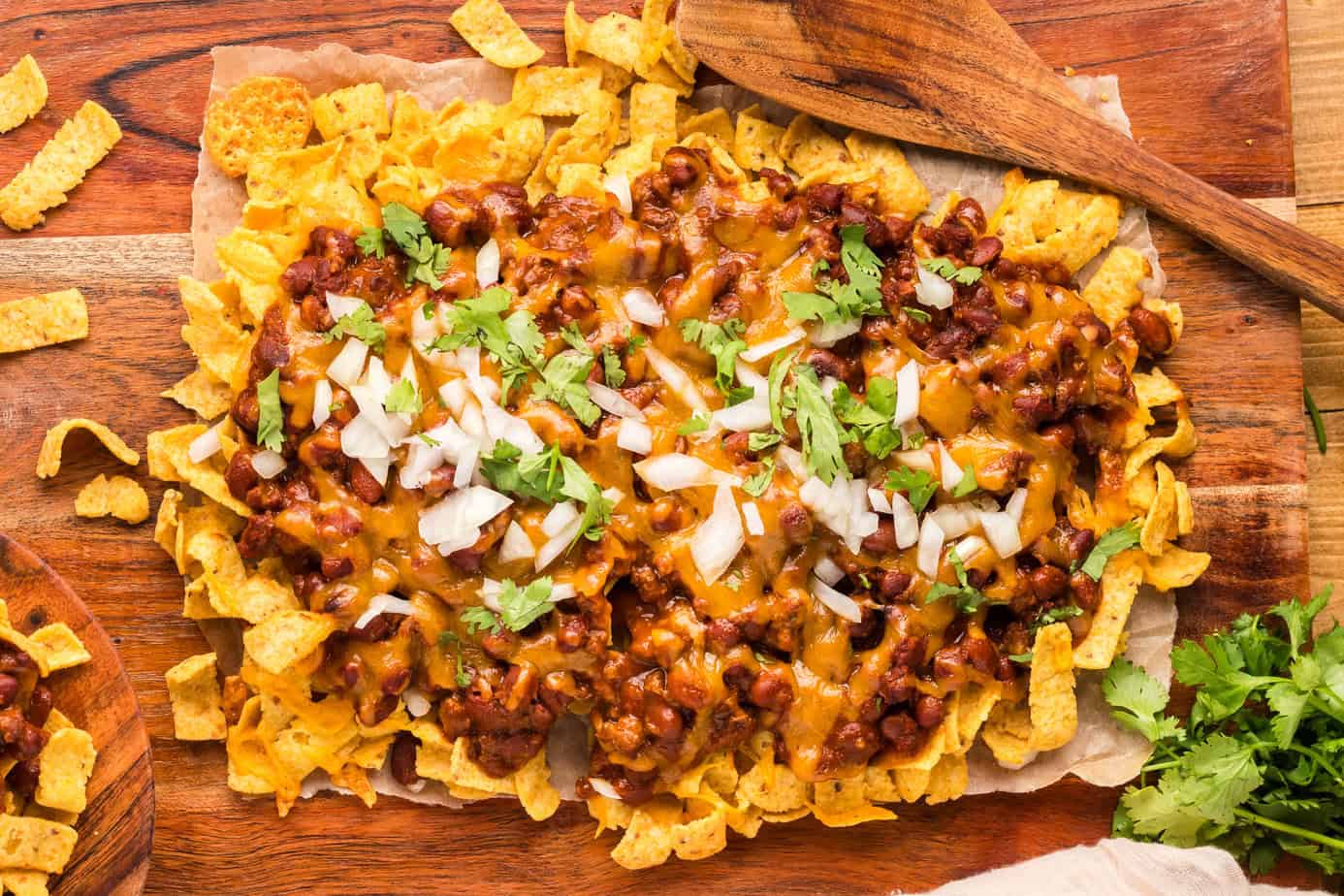 A sheet of corn chips topped with chili, melted cheese, chopped onions, and cilantro, served on parchment paper with a wooden spoon on the side.