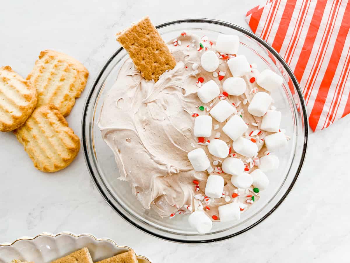 A glass bowl of creamy dip topped with mini marshmallows and crushed peppermint, with a graham cracker stick, next to striped cookies and a red-striped napkin.