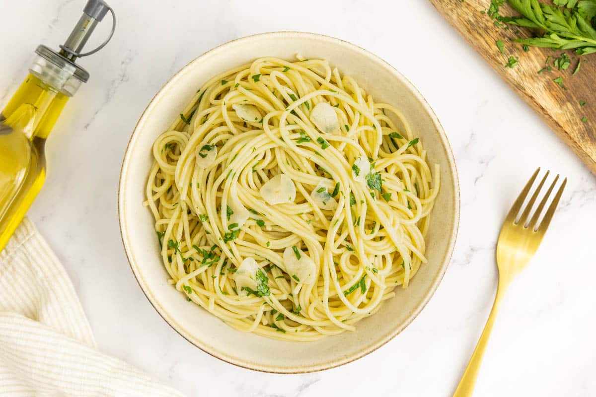 A bowl of spaghetti aglio e olio garnished with chopped parsley, next to a fork, olive oil bottle, and a wooden board with parsley on a white surface.