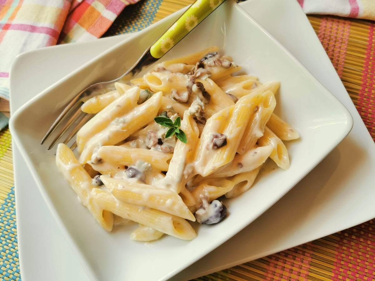 A square white plate with creamy penne pasta and mushrooms, garnished with a small sprig of herbs, with a fork resting on the plate.