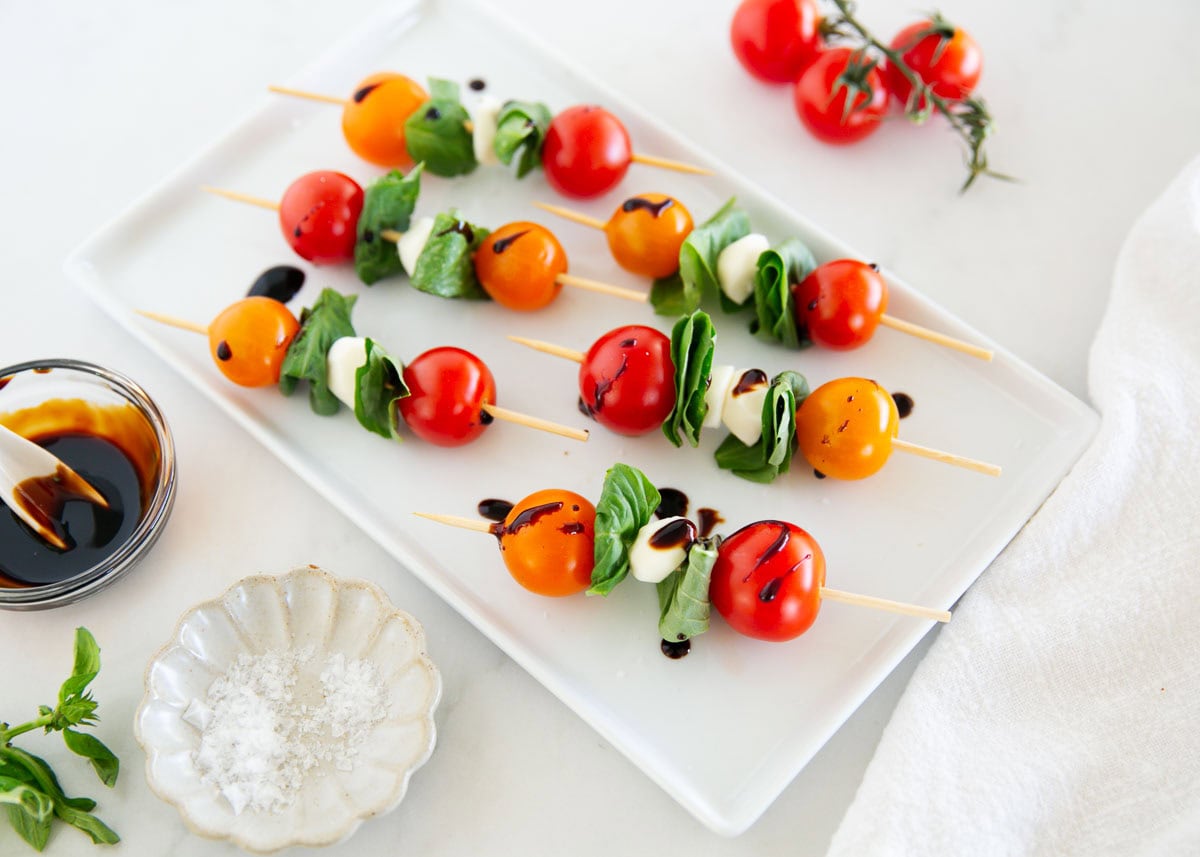 Skewers with cherry tomatoes, mozzarella balls, and basil leaves drizzled with balsamic glaze on a white plate, with salt, basil, and tomatoes nearby.