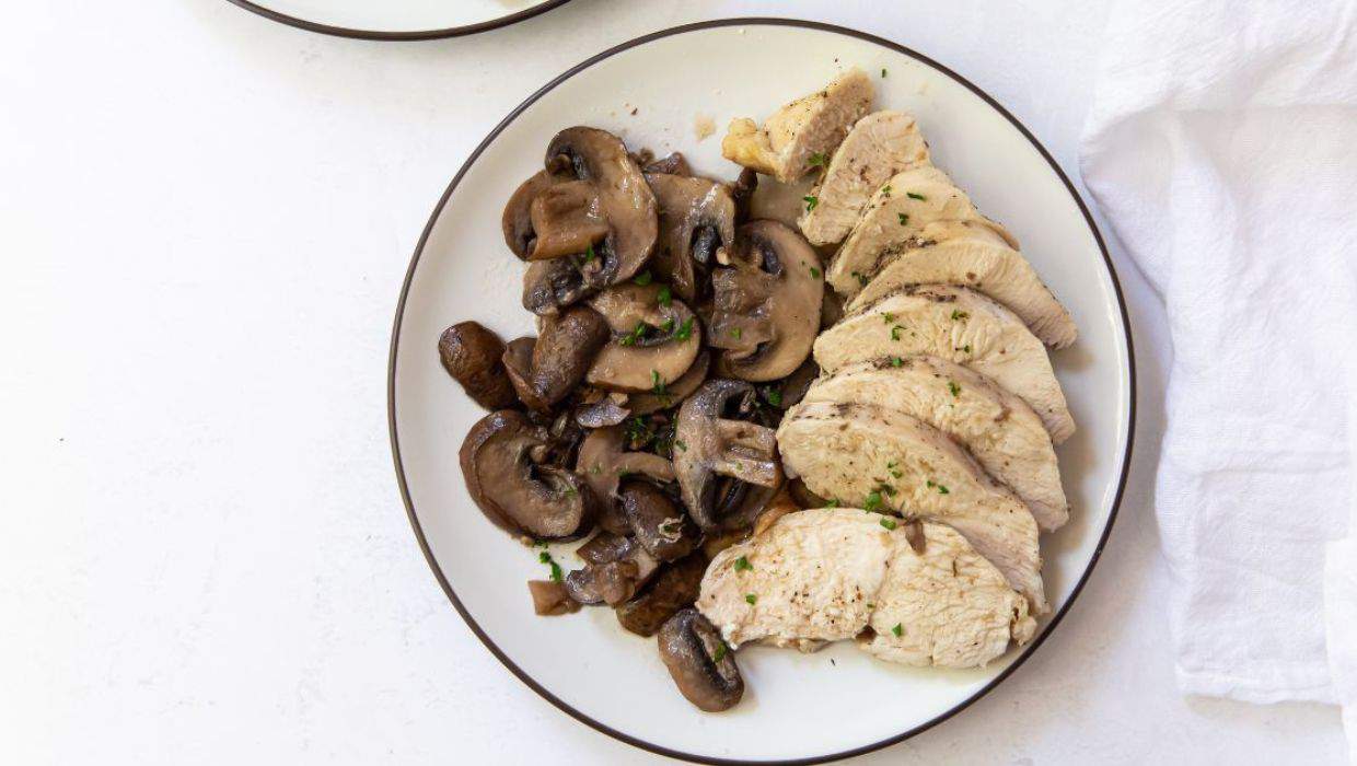 A white plate with sliced cooked chicken breast and sautรฉed mushrooms, garnished with herbs.