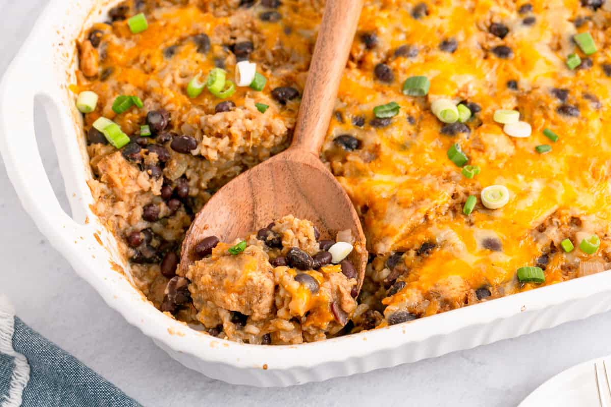 A baked casserole dish filled with a cheesy rice, black bean, and vegetable mixture, topped with melted cheese and chopped green onions. A wooden spoon holds a serving.