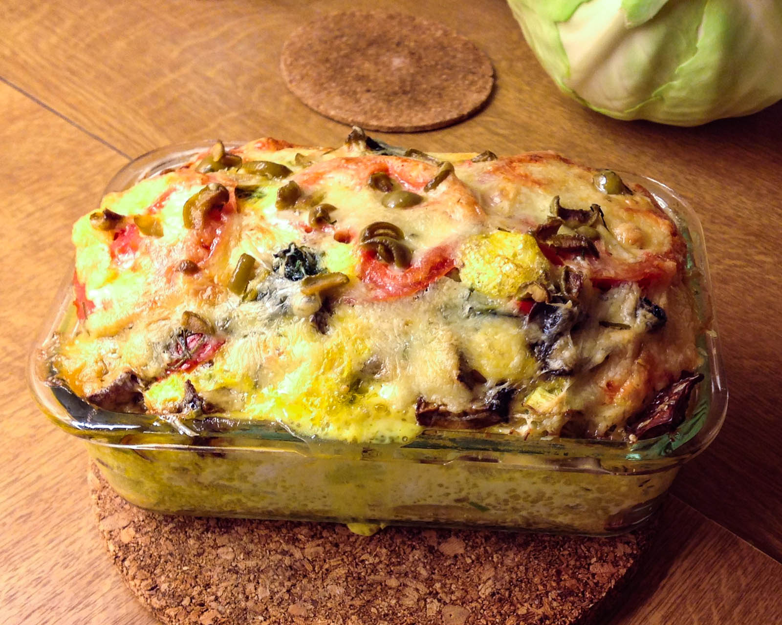 A rectangular glass dish filled with a baked vegetable casserole topped with melted cheese, placed on a cork trivet on a wooden table.