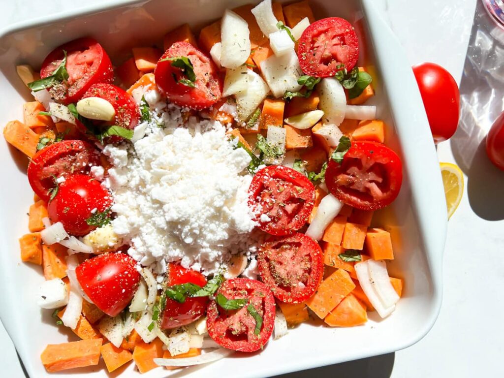 Chopped sweet potatoes, onions, tomatoes, garlic cloves, fresh basil, and crumbled cheese are arranged in a white baking dish, ready for roasting.