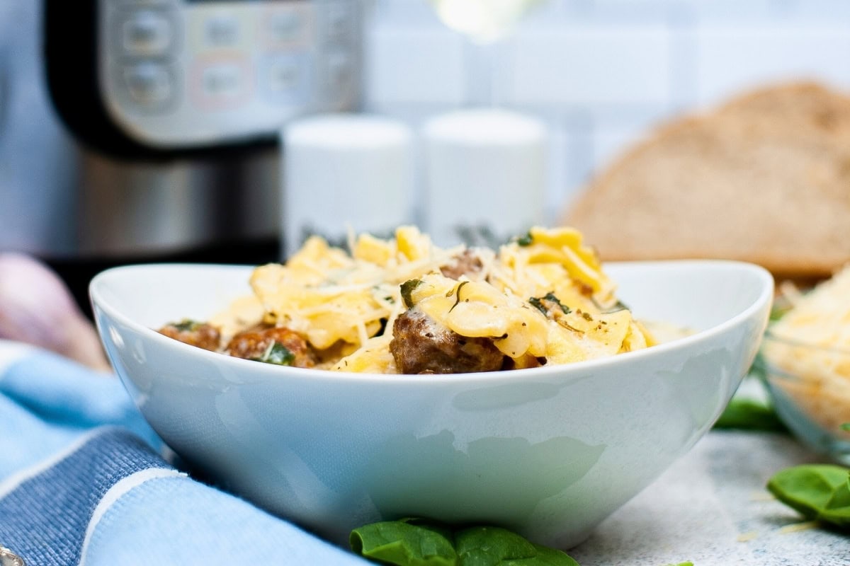 A white bowl filled with pasta, meatballs, and grated cheese, with a bread slice and kitchen appliances in the background.