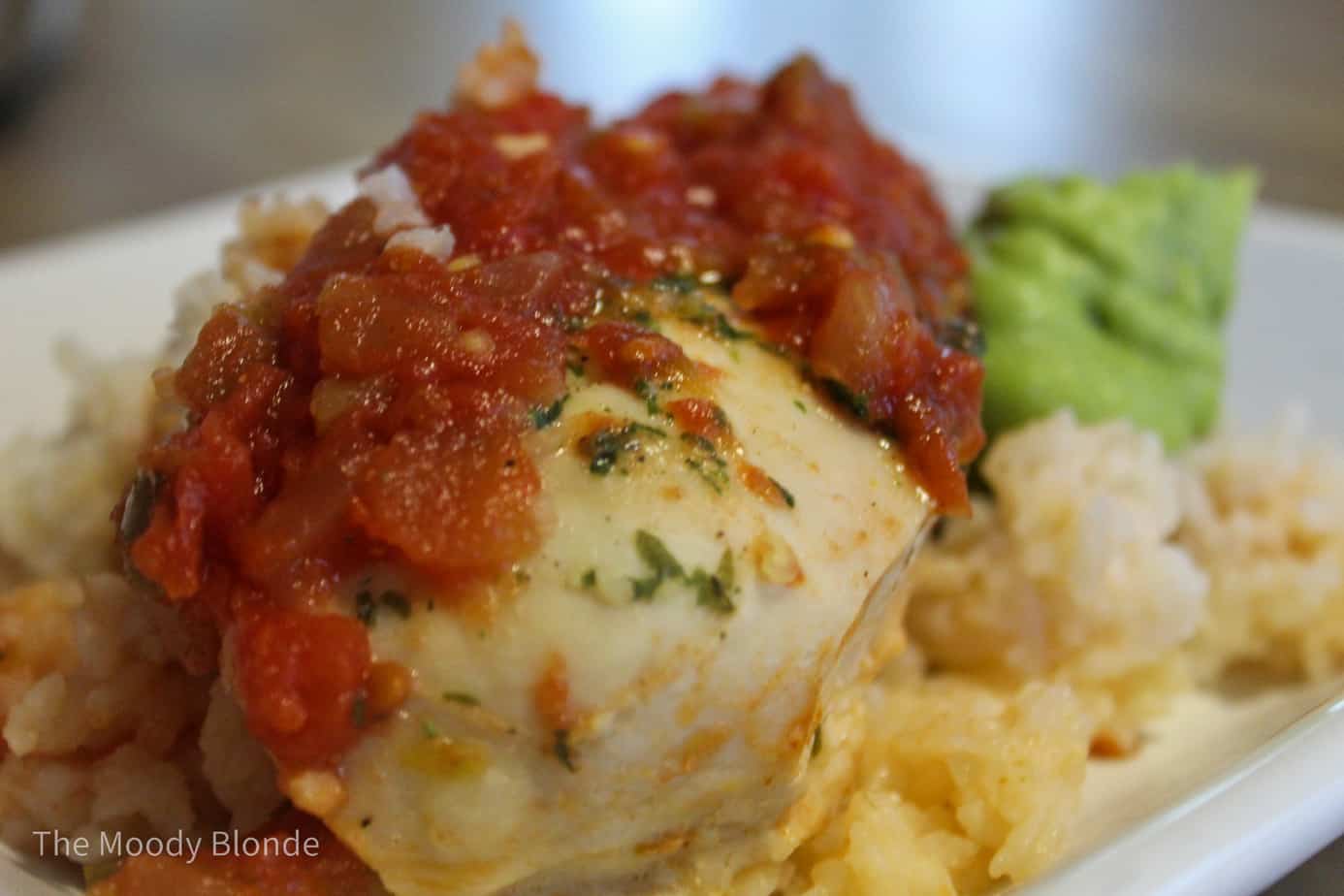 A piece of baked chicken breast topped with tomato sauce is served on a bed of rice with a small portion of avocado on a white plate.