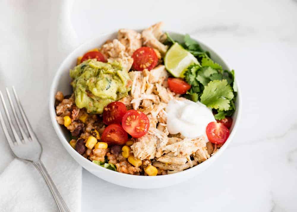 A white bowl containing shredded chicken, corn, black beans, cherry tomatoes, guacamole, sour cream, cilantro, and a lime wedge on a white marble surface with a fork nearby.