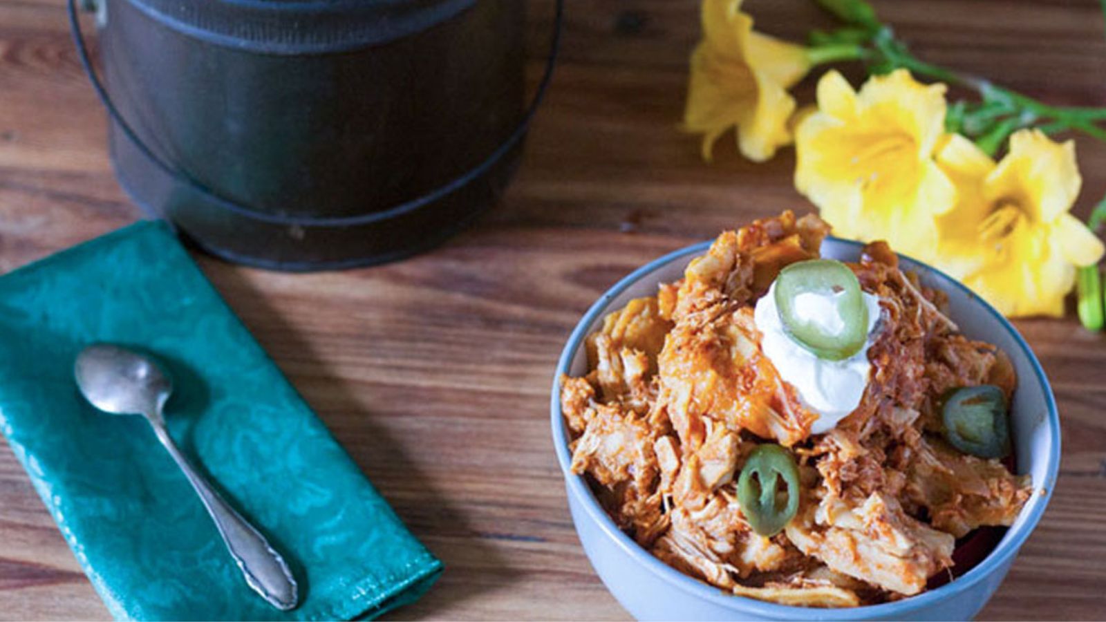 Bowl of shredded chicken topped with sour cream and jalapeรฑo slices, placed on a wooden table near a teal napkin with a spoon and yellow flowers.
