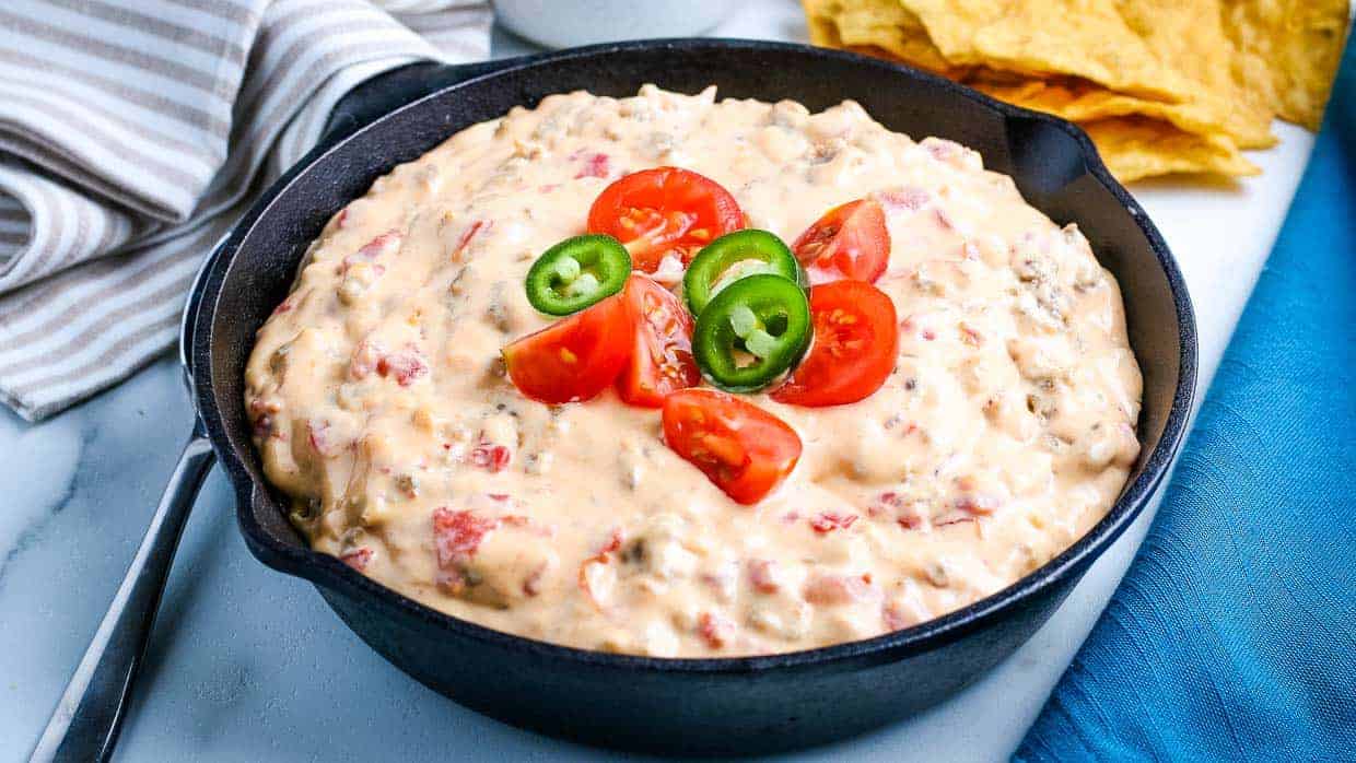A cast iron skillet filled with cheesy dip topped with sliced cherry tomatoes and jalapeรฑos, with tortilla chips on the side.