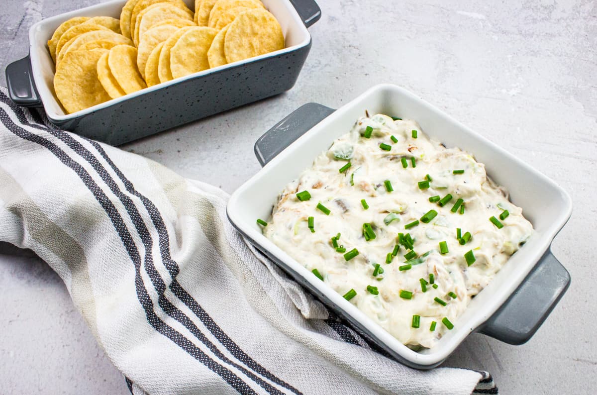 A square dish of creamy dip topped with chopped chives sits next to a container of round crackers on a white surface with a striped kitchen towel.
