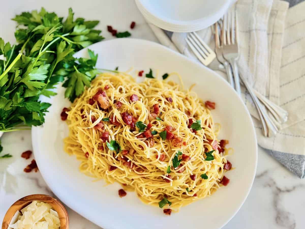 A white platter of pasta garnished with parsley and diced bacon, placed on a marble surface beside a bunch of fresh parsley, grated cheese, and stacked plates with utensils.