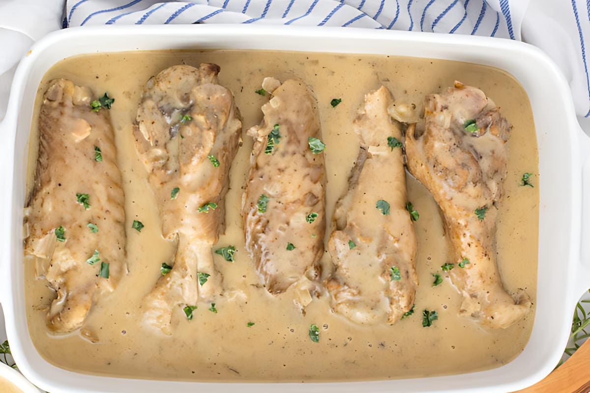 Four cooked chicken drumsticks in a white baking dish, covered in creamy gravy and garnished with chopped parsley.