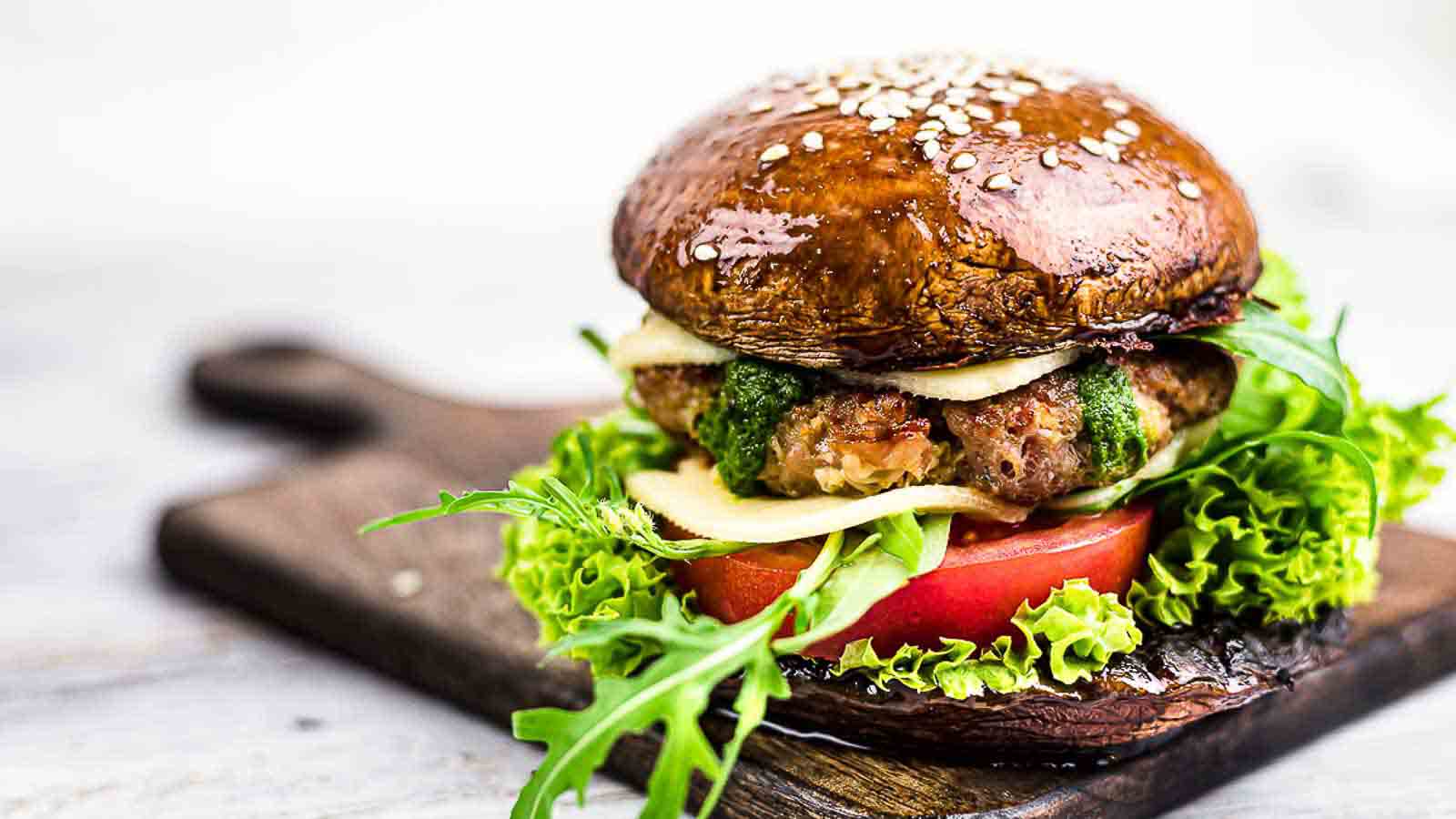 A burger with a portobello mushroom cap for a bun, filled with lettuce, tomato, cheese, arugula, pesto, and a patty, served on a wooden board.