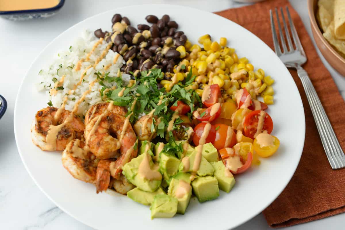 A white plate with grilled shrimp, diced onion, black beans with corn, yellow corn, halved cherry tomatoes, avocado chunks, and chopped cilantro, next to a fork and napkin.