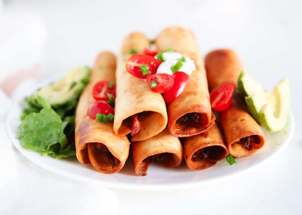 A plate of crispy taquitos topped with sour cream, sliced cherry tomatoes, and green onions, served with lettuce and avocado slices on the side.