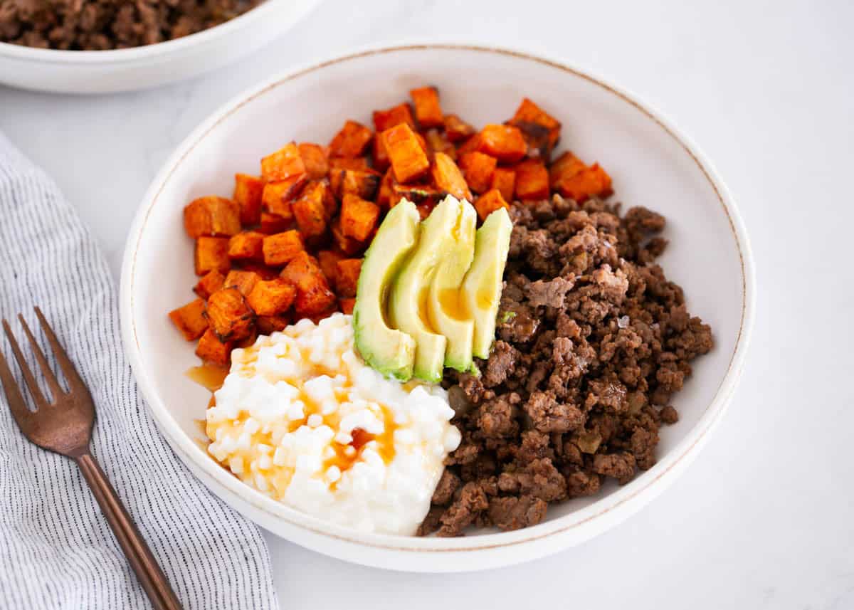 A bowl with ground beef, roasted sweet potatoes, sliced avocado, and cottage cheese, placed on a white surface next to a fork and a striped napkin.