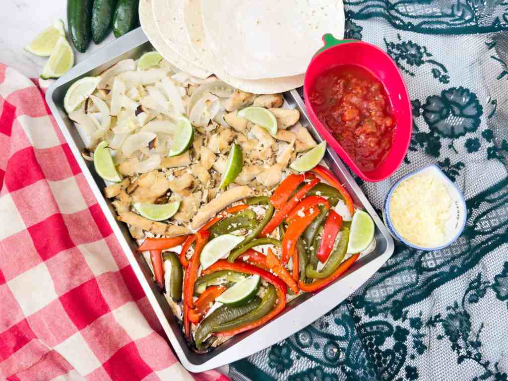 A tray with cooked chicken, sliced onions, bell peppers, and lime wedges, next to tortillas, a bowl of salsa, and a small bowl of shredded cheese on a patterned tablecloth.