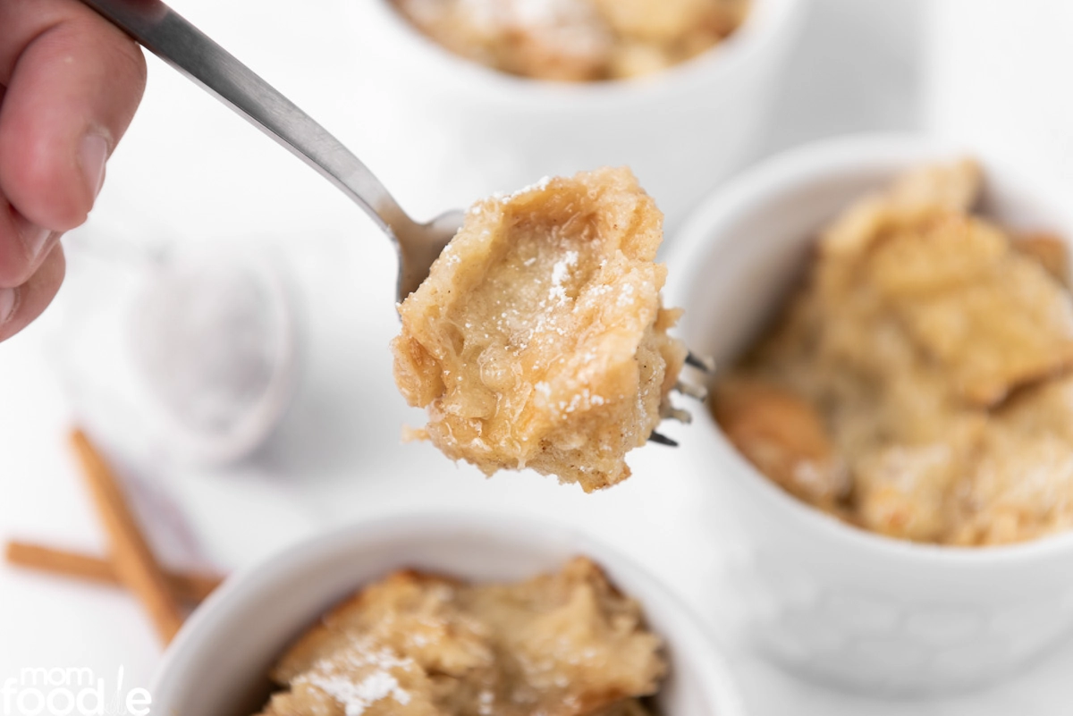 A hand holds a fork with a bite of bread pudding, with ramekins of bread pudding and a dusting of powdered sugar visible in the background.